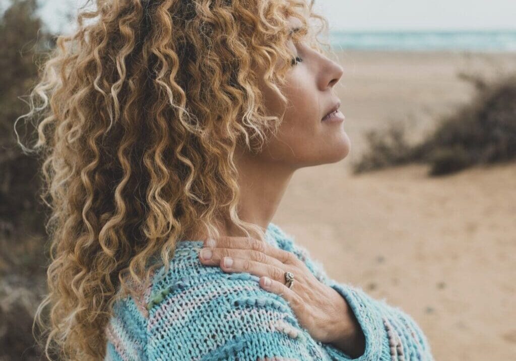 Woman with curly hair in a sweater, standing on a beach with eyes closed.
